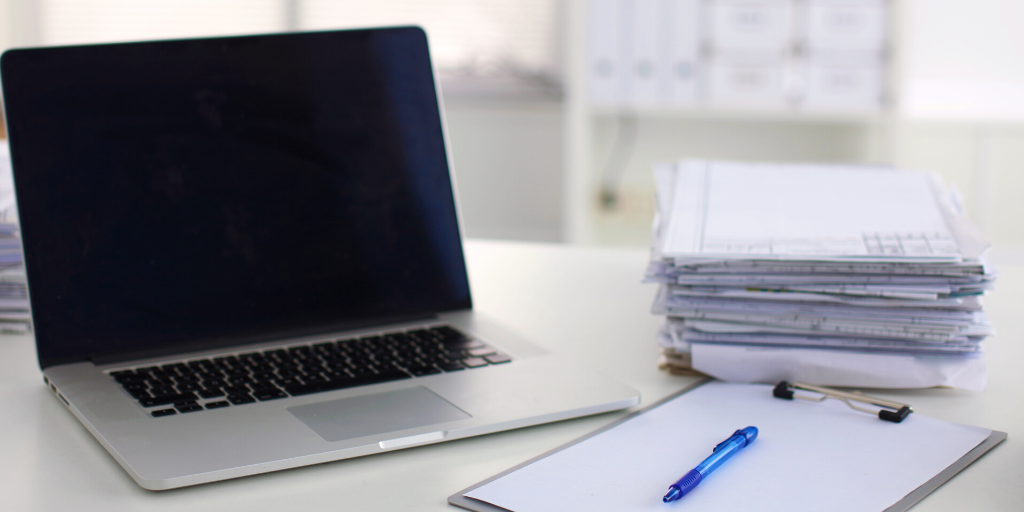 laptop next to a pile of paperwork and a pen