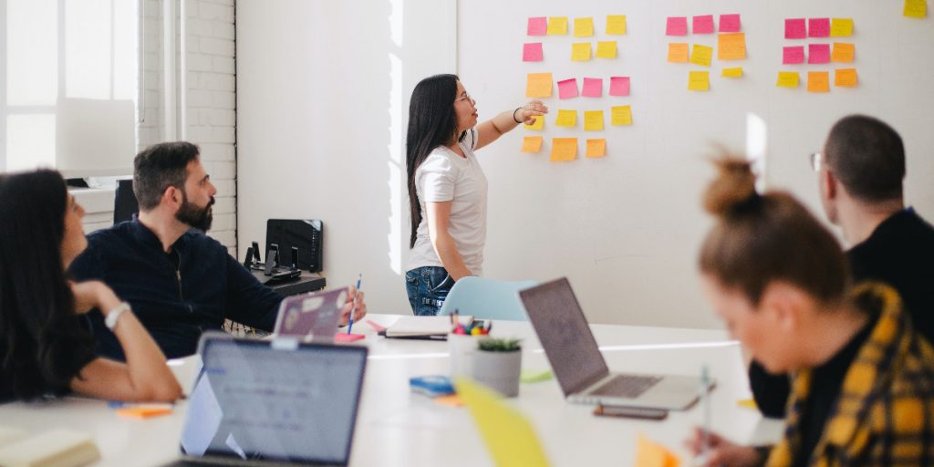 woman pointing at whiteboard and post its in front of classroom with laptops