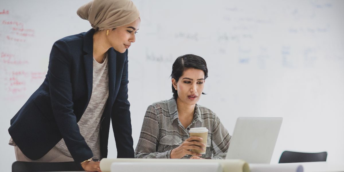 two women discuss something on a laptop in front of a whiteboard