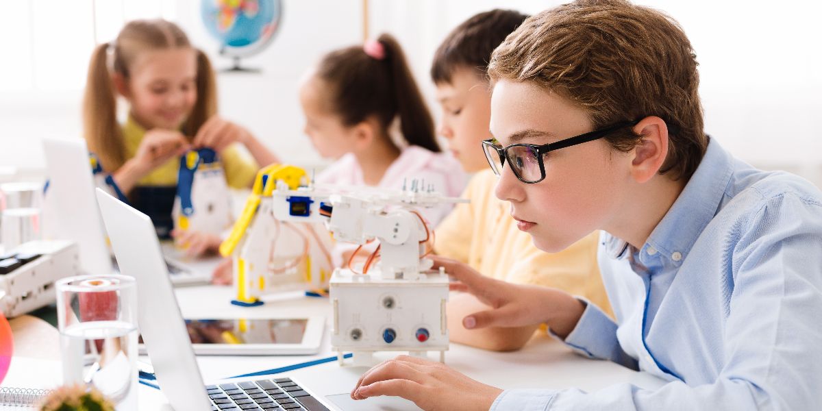 young boy on laptop assembling robot with classmates in background