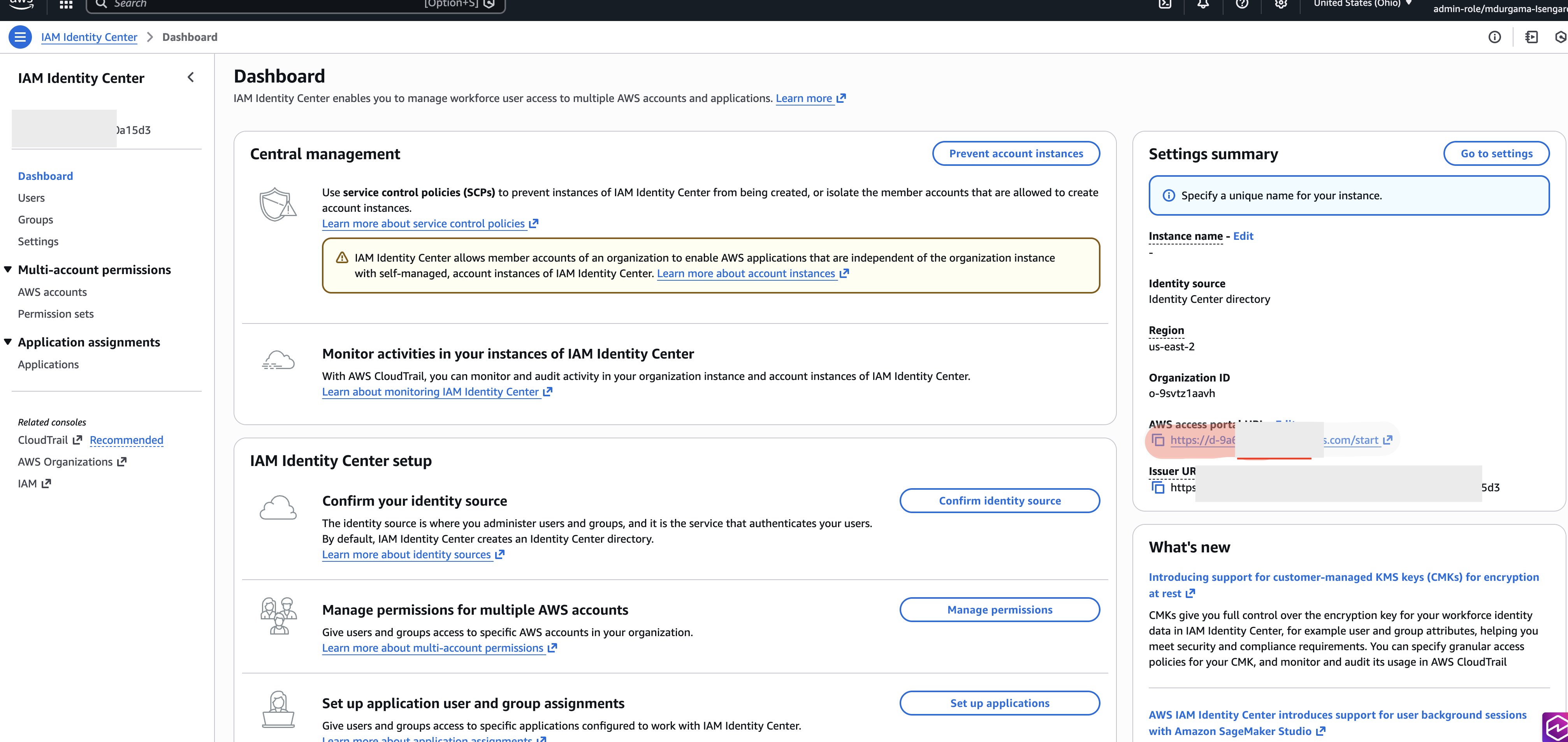 AWS IAM Identity Center Dashboard displaying left navigation menu with Dashboard, Users, Groups, Settings, Multi-account permissions (AWS accounts, Permission sets), and Application assignments sections; central management panel showing service control policies guidance with yellow warning banner about member account instances and CloudTrail monitoring section; IAM Identity Center setup area with three action cards for confirming identity source, managing multi-account permissions, and setting up application assignments; right panel Settings summary showing Identity Center directory as identity source, us-east-2 region, organization ID o-9svtz1aavh, AWS access portal URL, and issuer URL; What's new section highlighting customer-managed KMS keys support and Amazon SageMaker Studio user background sessions; Related consoles links to CloudTrail, AWS Organizations, and IAM.