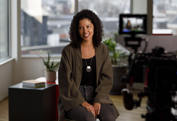 A behind the scenes photo of a smiling woman with medium skin tone and curly dark hair being interviewed on camera.