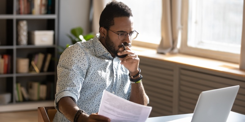 Focused african businessman holding documents looking at laptop doing research