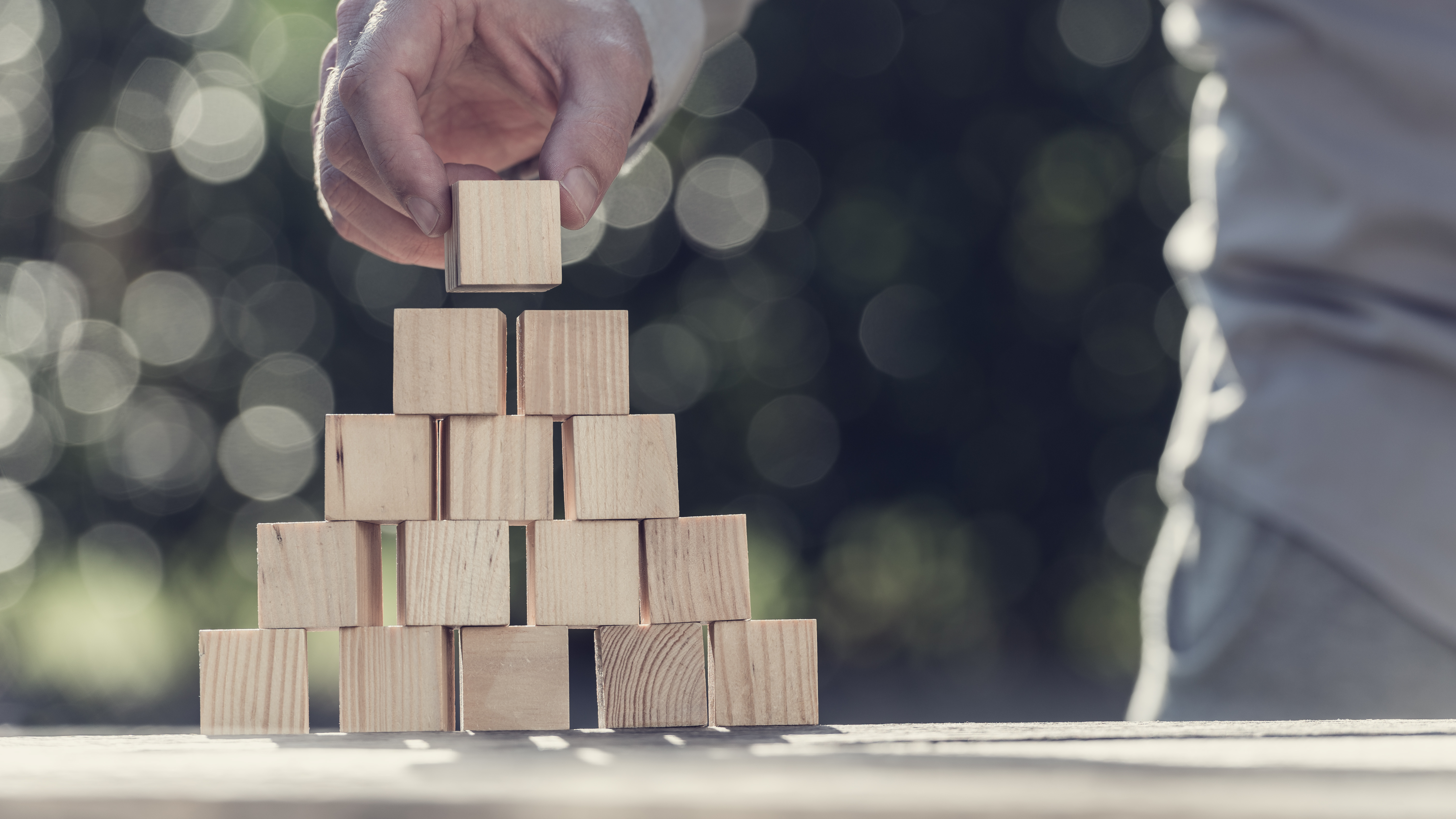 Retro toned image of man building a pyramid with blank wooden blocks outdoors against sparkling bokeh.