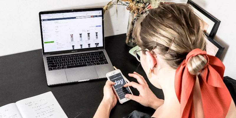 woman looks at her phone and shops online with a laptop in front of her