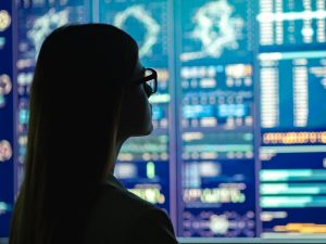 Profile of a woman in glasses reviewing a backlit display of several analytical dashboards 