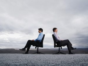 Two men sitting in office chairs outdoors with their backs against one another