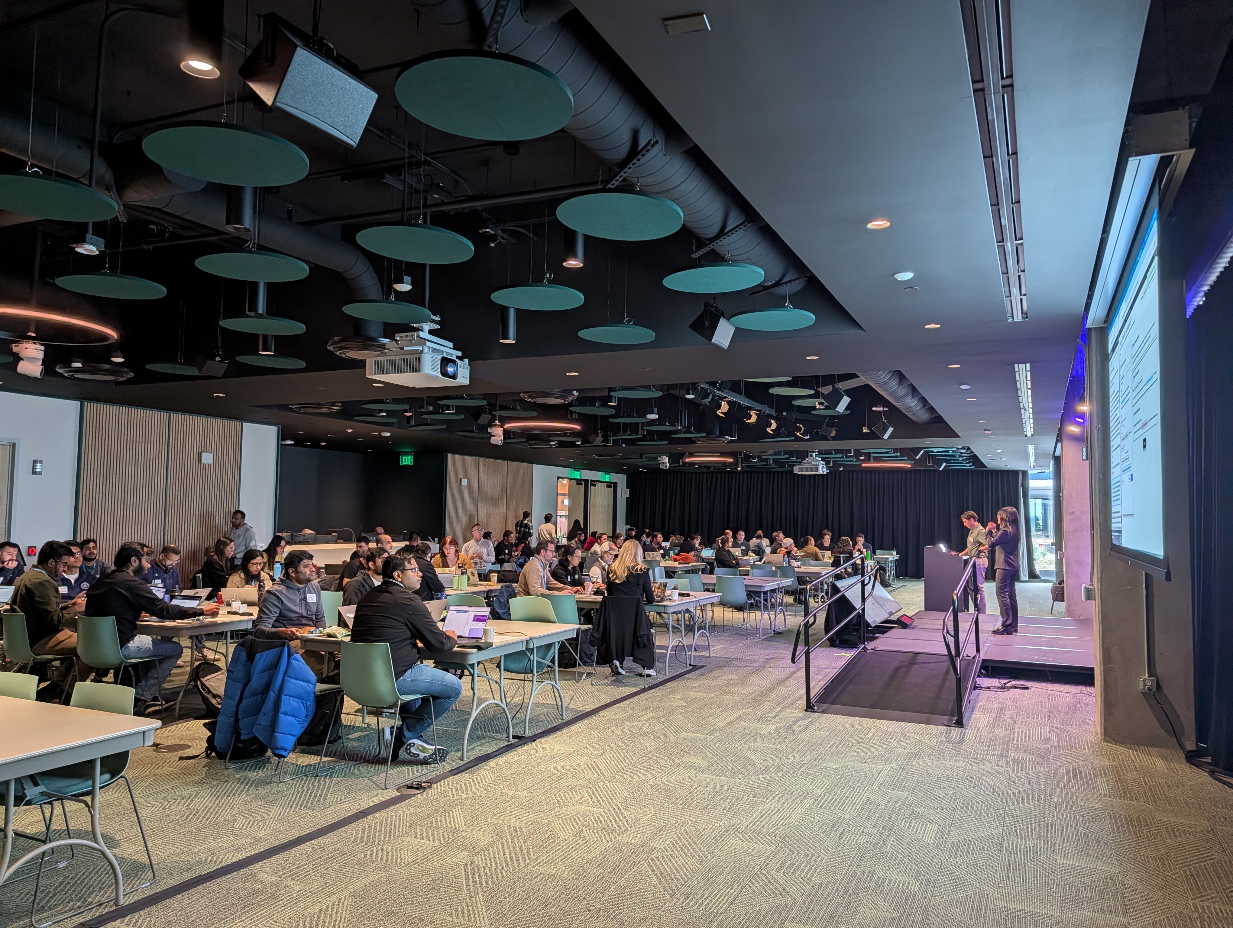 Modern conference hall with attendees seated at tables, listening to a presentation by two speakers on stage. Large screens display slides, and the room features circular acoustic panels and exposed ductwork, indicating a professional or academic event setting.