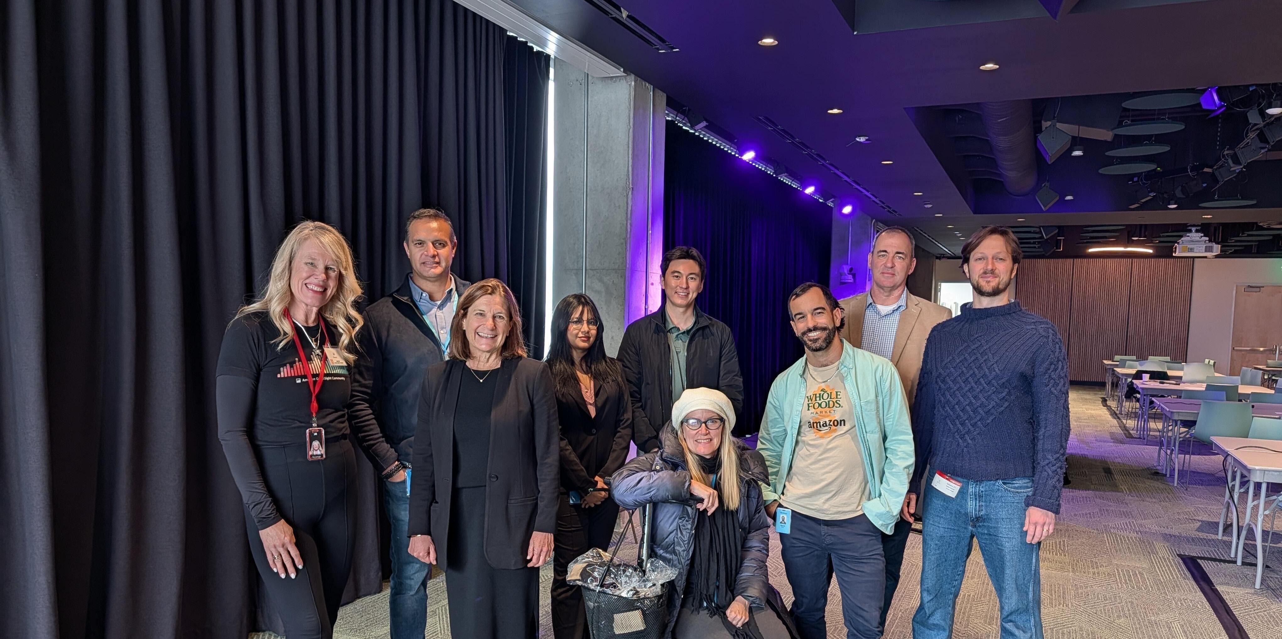 A diverse group of eight people posing in a modern event venue with purple lighting, black curtains, and a stage area, dressed in business casual attire, suggesting a professional or semi-formal gathering.