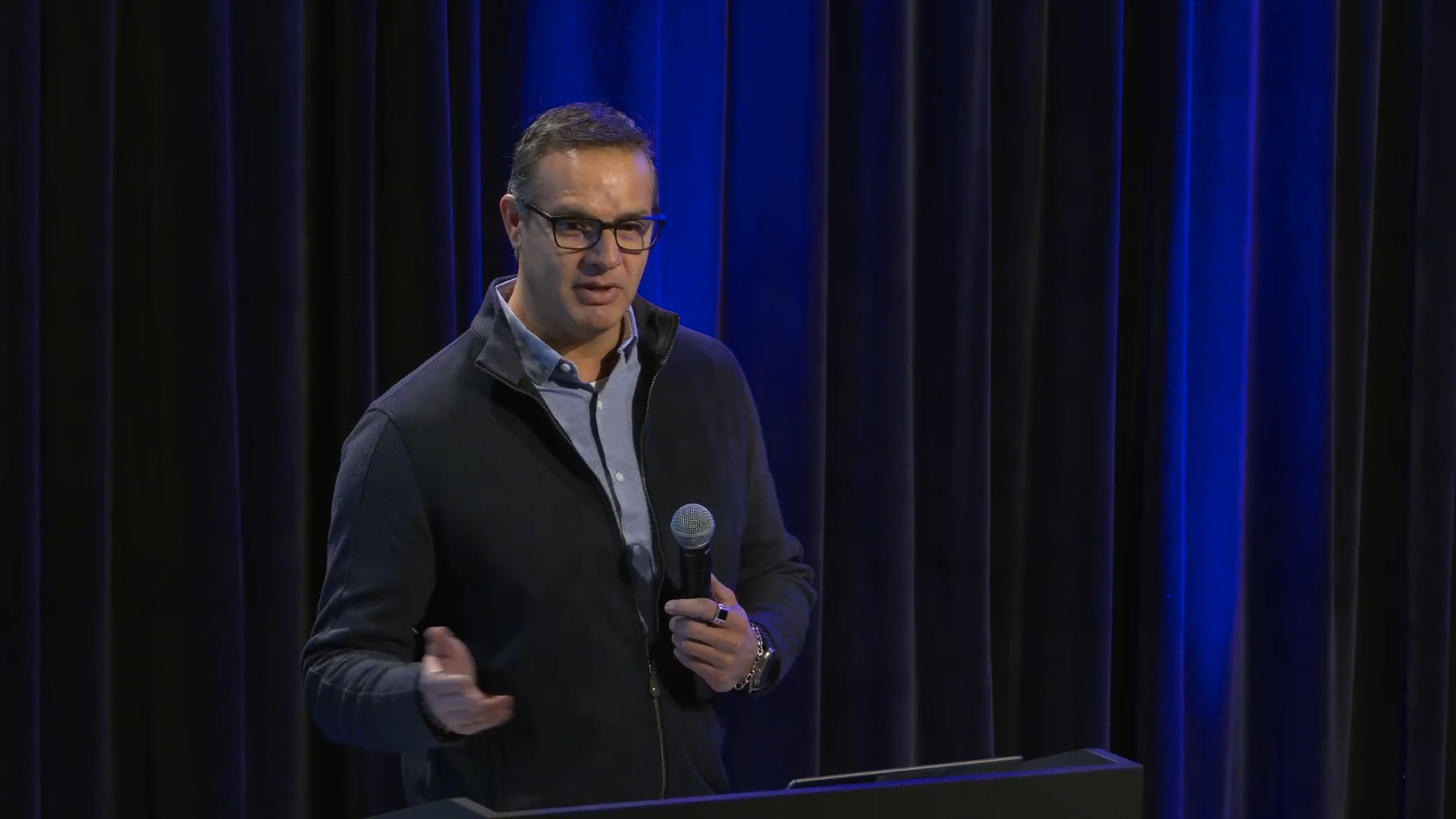 Man with glasses wearing a dark jacket and light-colored shirt stands in front of a dark blue curtain backdrop, holding a microphone and gesturing with his hand while speaking at a professional event.