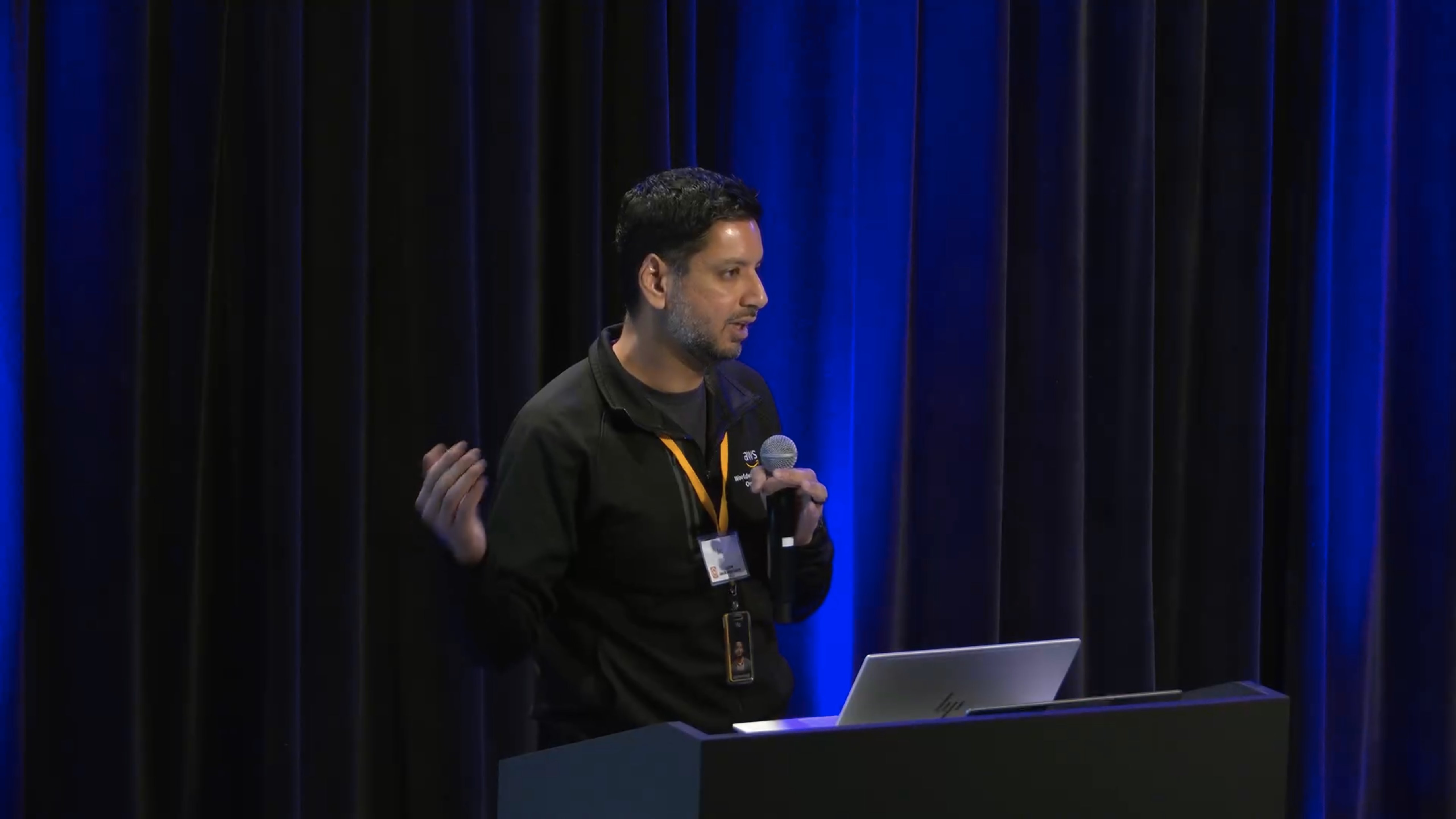 Man in a black hoodie with an orange lanyard and badge, holding a microphone and speaking in front of a laptop on a stand, against a dark blue curtain backdrop.