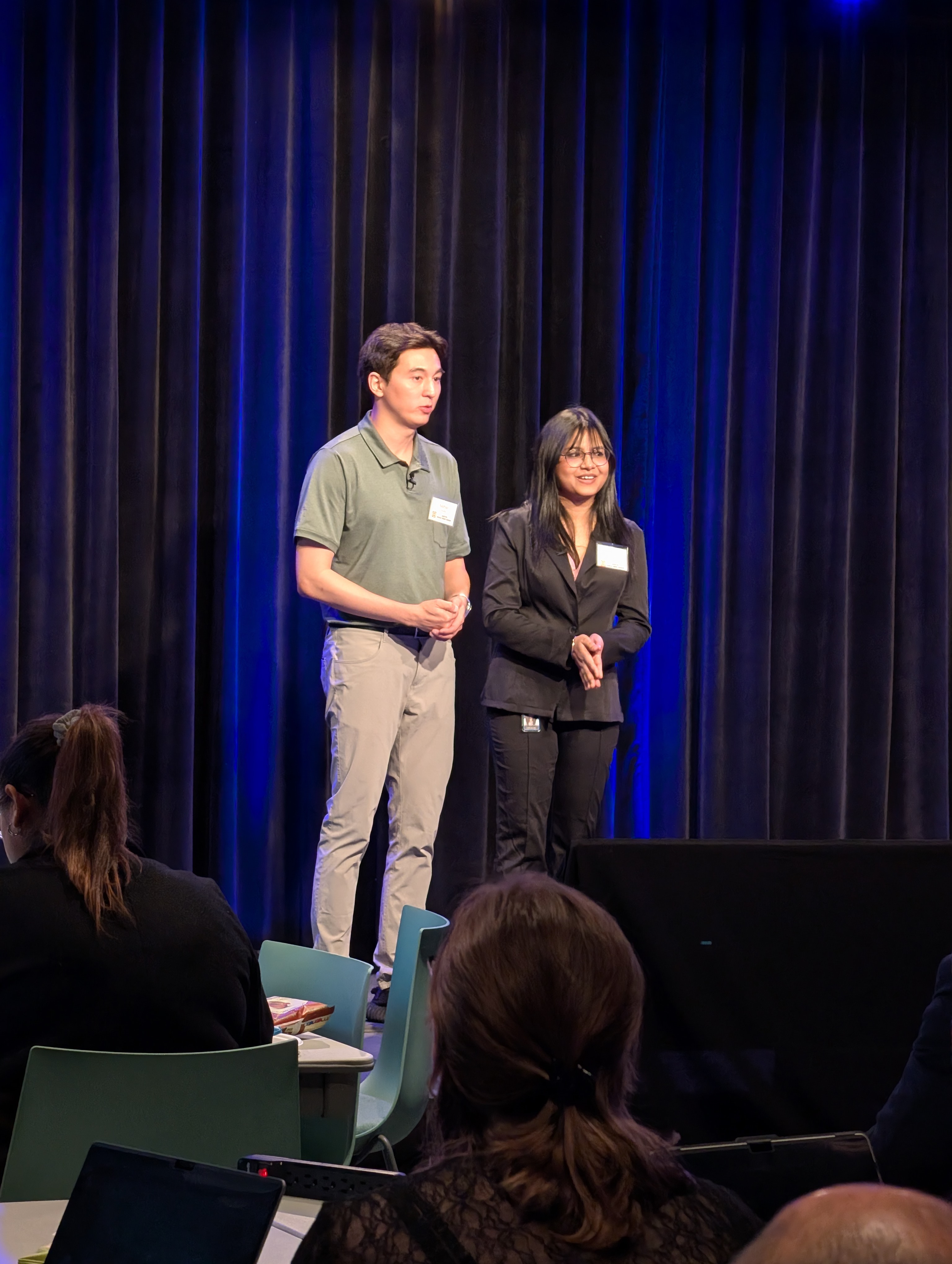 Two presenters, one in a green shirt and beige pants, the other in a black suit, speaking on stage in front of a dark blue curtain with an audience seated before them, suggesting a formal conference or seminar setting.