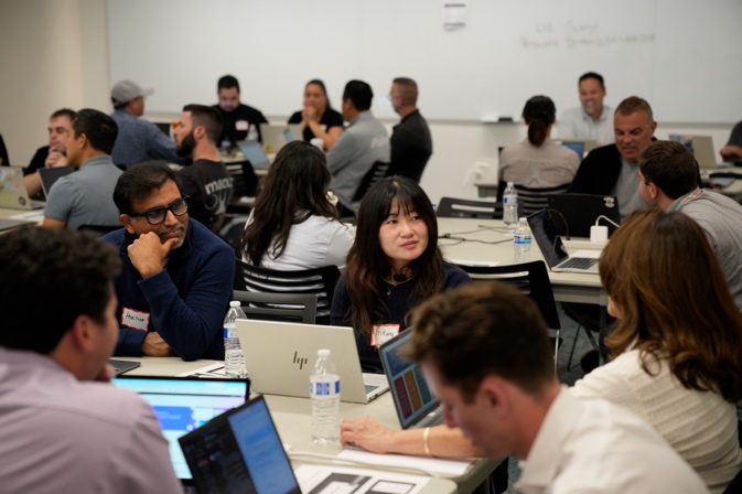 Approximately 15-20 attendees seated around tables with laptops and notebooks during an interactive workshop session at the Amazon Quick User Group meetup. Participants are engaged in discussion and collaboration.