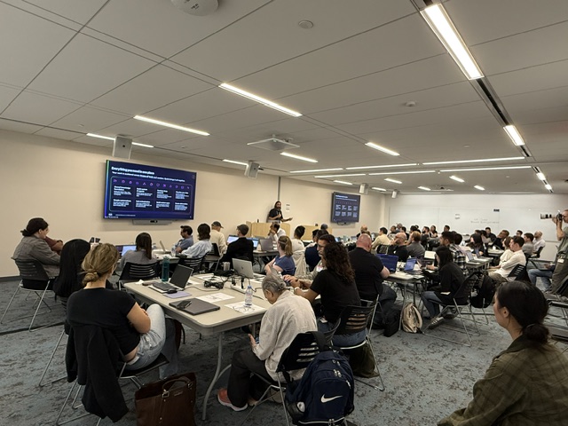 A large classroom filled with approximately 40-50 attendees seated at rows of tables with laptops during the Amazon Quick User Group meetup. Blue display screens are visible at the front of the room, and a speaker stands near the front.