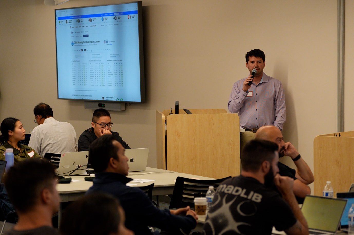 A presenter in a lavender button-up shirt speaking at a wooden podium with a microphone during the Amazon Quick User Group meetup. A monitor behind him displays a data tracking dashboard. Audience members are seated at tables with laptops, listening attentively.