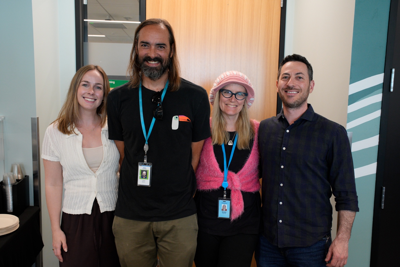 Four event participants posing together for a group photo in a modern office space during the Amazon Quick User Group meetup. Two of the four are wearing blue lanyards with ID badges, suggesting they are organizers or staff.