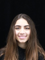 Professional headshot portrait of young woman with long brown hair smiling at camera against dark background, suitable for identification or professional profile