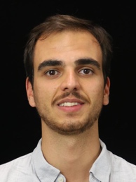 Professional headshot portrait of male subject with dark hair and facial stubble wearing white collared shirt against plain black background