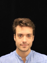 Professional headshot portrait of young man with light brown hair wearing light blue button-up shirt photographed against black background in neutral frontal pose