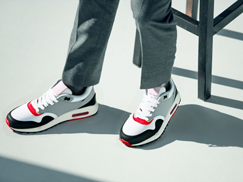 Product photograph of white, gray, red, and black color-blocked athletic sneakers with white laces worn with gray pants against minimalist white background with geometric shadows