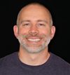 Professional headshot portrait of a man with a beard smiling against a dark background, wearing a dark short-sleeved shirt.