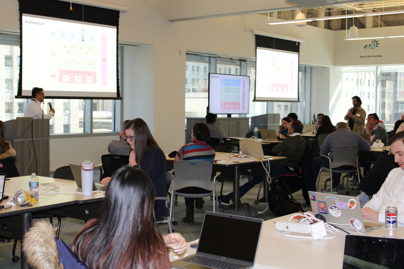 Attendees working on laptops at tables during a session, with an architecture slide on screen