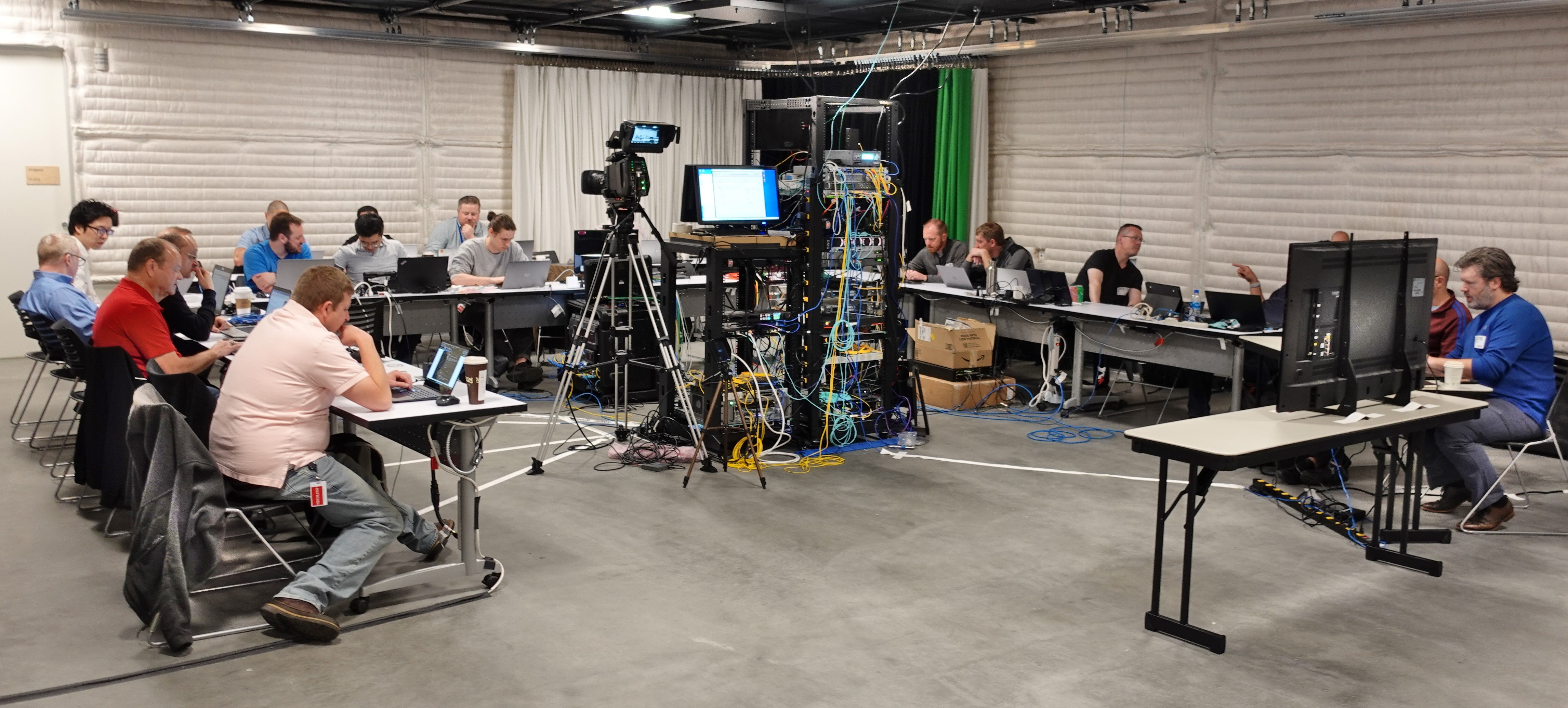 Participants at the JPEG XS interoperability workshop sit at a U-shaped set of desks surrounding two racks of JPEG XS equipment as well as a broadcast video camera.