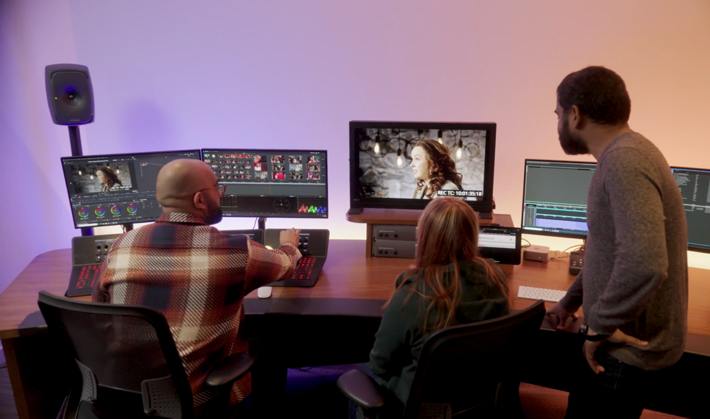 Three people collaborate in a video editing studio, reviewing footage on multiple monitors displaying color grading tools and editing timelines. One person operates the editing console, while the others observe and discuss the scene on the central screen showing a woman being filmed.